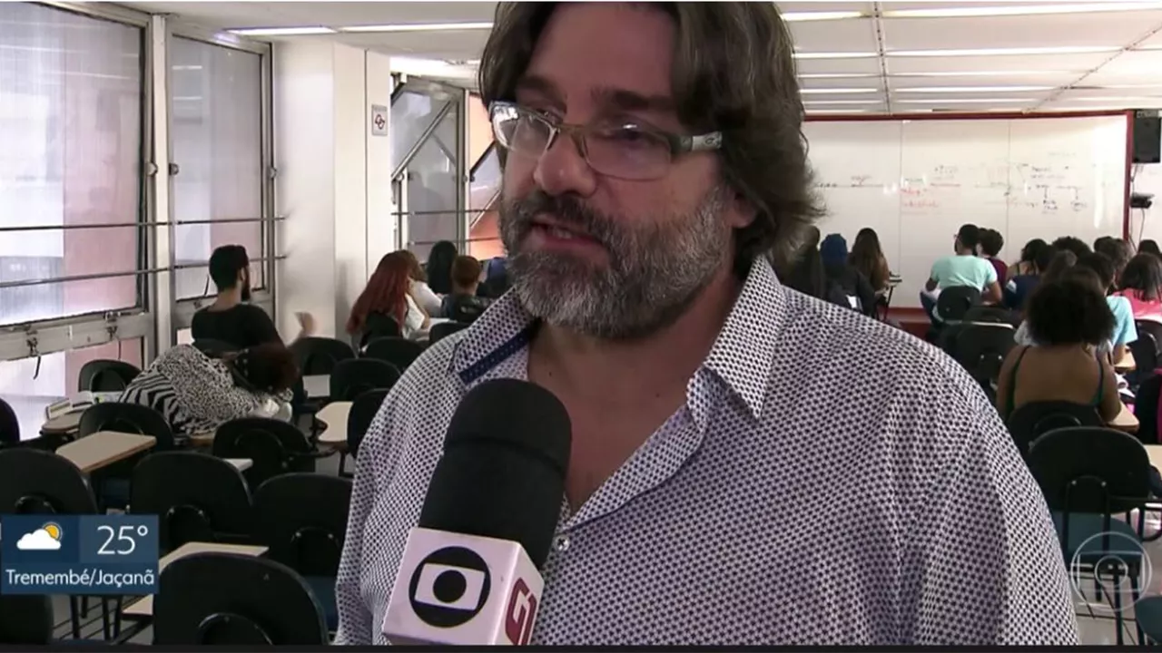 Sala de aula vista do fundo onde professor aparece em primeiro plano dando entrevista e seus alunos aparecem no plano de fundo fazendo atividade