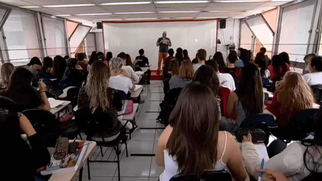 Sala de aula vista do fundo, onde alunos prestam atenção a professor, que lê um papel na frente da sala